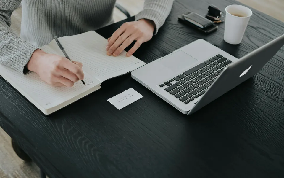 O persoană la birou notează date într-un carnet, lângă un laptop deschis